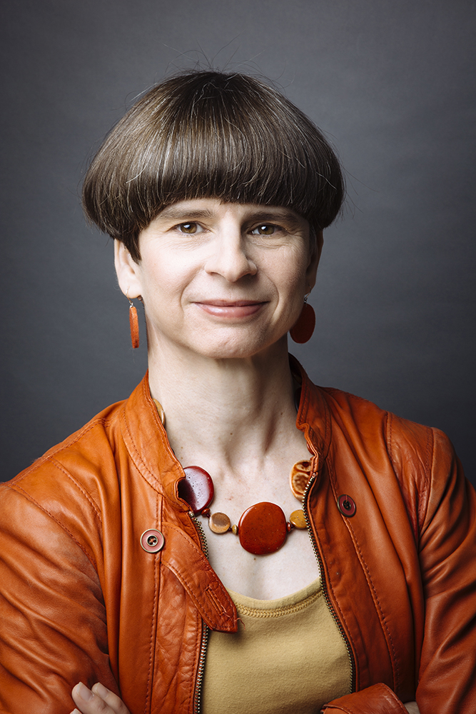 Portrait of Elizabeth Merritt, facing camera with arms crossed, in orange leather jacket and yellow blouse with orange disc-shaped earrings and pendant necklace, against a mottled gray background.
